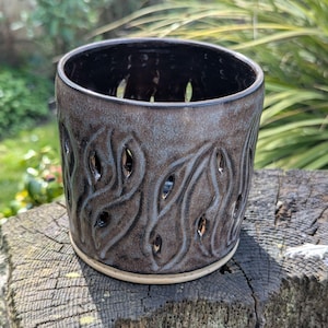 May include: A handcrafted ceramic candle holder with a dark brown glaze and a textured leaf-like pattern. The cylindrical holder features cut-out designs and a light-colored base. The candle holder is approximately 10 cm tall.