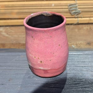 May include: A small, pink ceramic cup with a speckled glaze. The cup has a rounded bottom and a slightly flared rim.