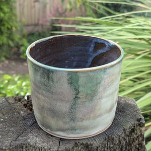 May include: A handmade ceramic tumbler with a speckled, muted green and grey exterior. The interior is a dark, glossy brown with hints of blue. The rim has a light brown accent. The tumbler is cylindrical and sits on a wooden surface.