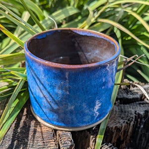 May include: A handcrafted ceramic mug with a striking blue glaze. The mug features a cylindrical form with a gently widening rim and an unglazed base. The interior is a dark brown hue.