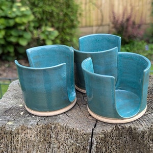 May include: Four teal ceramic spoon rests with a curved design. The spoon rests have a glossy glaze and sit on an unglazed base. The background shows green foliage and a wooden surface.