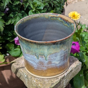 May include: A cylindrical ceramic planter featuring a reactive glaze with blended layers of earthy brown, soft blue, and sage green. The rim displays a darker, toasted brown tone with subtle drips cascading down the exterior surface of the vessel.