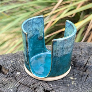 May include: A cylindrical ceramic sponge holder featuring a deep teal and turquoise glaze with a smooth, glossy finish. The vessel has a large U-shaped cutout on one side and a flat, unglazed beige base, showcasing a reactive glaze effect.