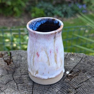 May include: A white ceramic vase with a light pink and brown glaze. The vase has a rounded bottom and a wide opening at the top. The inside of the vase is black.