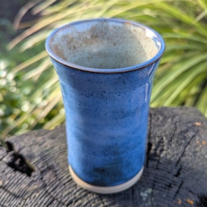 May include: A handmade ceramic tumbler with a speckled blue glaze. The interior has a light tan colour, and the rim is a darker blue. The tumbler is cylindrical with a slightly flared top and sits on a small, unglazed base.