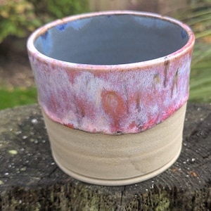 May include: A handmade ceramic cup with a unique glaze. The cup features a light beige base, transitioning to a vibrant red and white speckled midsection, and a blue interior. The cup is sitting on a wooden surface.