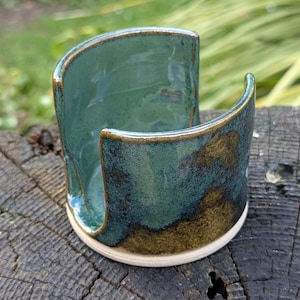 May include: A teal and brown glazed ceramic sponge holder with a curved design. The handmade holder has a glossy finish and a light beige base. It is displayed on a weathered wooden surface.