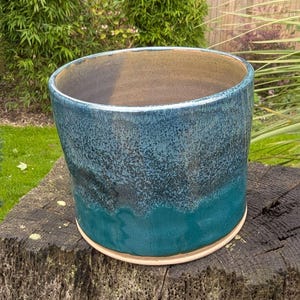 May include: A cylindrical ceramic planter featuring a reactive glaze with a gradient of deep teal at the base transitioning into a speckled blue and dark grey finish toward the rim, with a smooth, light beige interior and a flat, circular base.