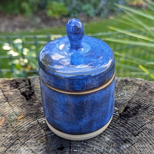 May include: A handmade, cobalt blue ceramic jar with a lid and a small knob. The jar is cylindrical with a light beige base and a thin, gold-coloured band around the middle. The lid fits snugly on top.