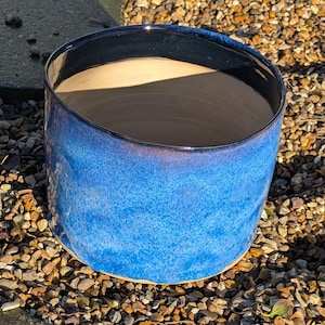 May include: A blue ceramic planter with a dark blue rim. The planter has a mottled blue glaze and is sitting on a bed of small stones.