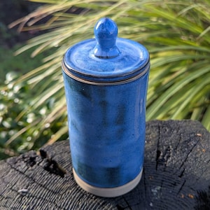 May include: A handmade, cobalt blue ceramic jar with a lid and a small knob. The jar has a cylindrical shape and a cream-coloured base. The lid fits snugly, and the jar is displayed on a dark, textured surface, with green foliage in the background.