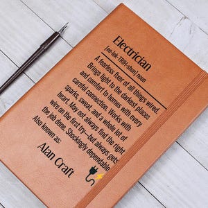 May include: A brown leather-bound notebook with the definition of "Electrician" printed in black. The text includes a description of an electrician's work and the words "Alan Craft" at the bottom. A black pen rests on the wooden surface.