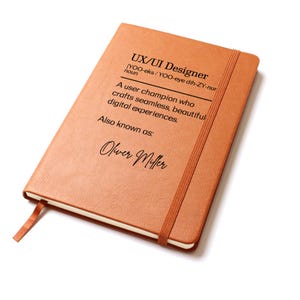 May include: A brown notebook with the text "UX/UI Designer" and the definition of the term. The notebook has a matching elastic band and a ribbon bookmark. The name "Oliver Miller" is also printed on the cover.