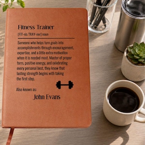May include: A tan leather-bound notebook featuring black printed text that defines a fitness trainer, including a dumbbell icon and the name John Evans. The cover displays the dictionary-style definition of the noun and its phonetic spelling.