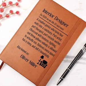 May include: A brown leather-bound notebook with the text "Interior Designer" and a definition. The definition describes an interior designer as a creative genius. The notebook also includes the name "Oliver Miller". A black pen rests on the side.