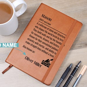 Könnte beinhalten: Ein braunes, ledergebundenes Notizbuch mit der Definition von "Mason" auf dem Einband sowie dem Namen "Oliver Miller". Eine weiße Kaffeetasse und drei Stifte liegen neben dem Notizbuch. Das Notizbuch hat ein Leseband.