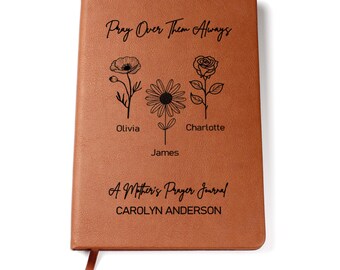 May include: A brown leather-bound journal with the text "Pray Over Them Always" and "A Mother's Prayer Journal" engraved on the front cover. The journal also features three flower illustrations and the names Olivia, James, and Charlotte.