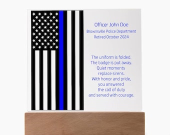 May include: A black and white American flag with a blue stripe down the right side. The text "Officer John Doe Brownsville Police Department Retired October 2024" is printed below the flag. A poem is printed below the text: "The uniform is folded. The badge is put away. Quiet moments replace sirens. With honour and pride, you answered the call of duty and served with courage."