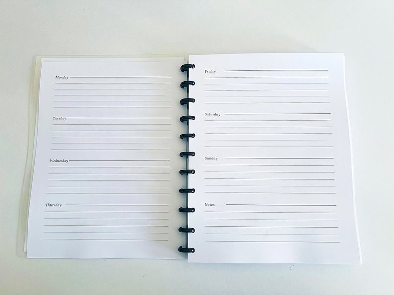 May include: A white planner with a black coil binding. The planner has a weekly layout with spaces for Monday through Sunday, as well as a notes section.
