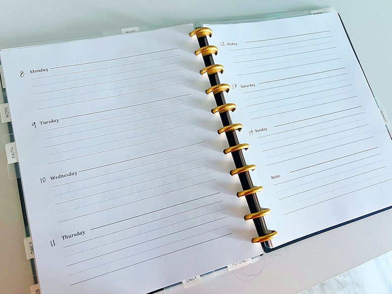 May include: A white planner with gold rings and tabs labeled "Daily", "Faith", "Work", and "Future". The planner has a weekly layout with space for notes and daily tasks. The days of the week are labeled "Monday", "Tuesday", "Wednesday", "Thursday", "Friday", "Saturday", and "Sunday".