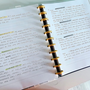 May include: A close-up of a white notebook with black ink. The notebook has a gold ring binder and is open to a page with handwritten notes. The notes include bible verses and scripture references. The page is dated with the days of the week and numbered.