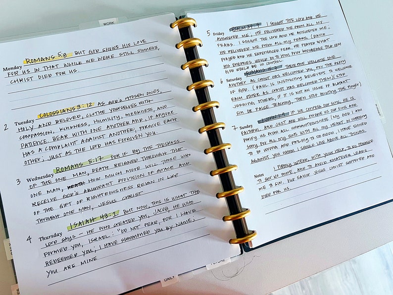 May include: A close-up of a white notebook with black ink. The notebook has a gold ring binder and is open to a page with handwritten notes. The notes include bible verses and scripture references. The page is dated with the days of the week and numbered.