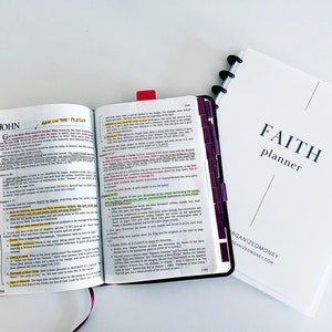 May include: An open Bible with the words "God in the Flesh" highlighted in yellow. The Bible is open to the book of John, chapter 1. The Bible is sitting on a white surface next to a white planner with the words "FAITH planner" printed on the front.
