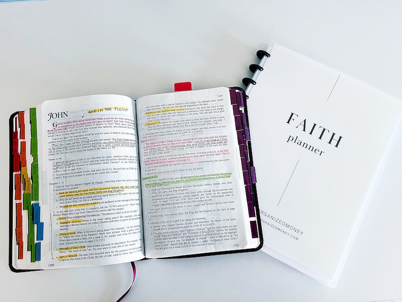 May include: An open Bible with the words "God in the Flesh" highlighted in yellow. The Bible is open to the book of John, chapter 1. The Bible is sitting on a white surface next to a white planner with the words "FAITH planner" printed on the front.