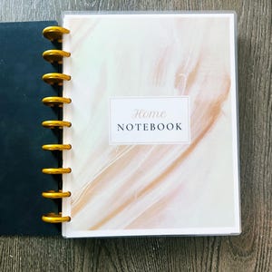 May include: A Home Notebook with a soft, abstract design in shades of peach and white. The notebook is bound with gold-colored rings and has the words "Home Notebook" printed on the cover.