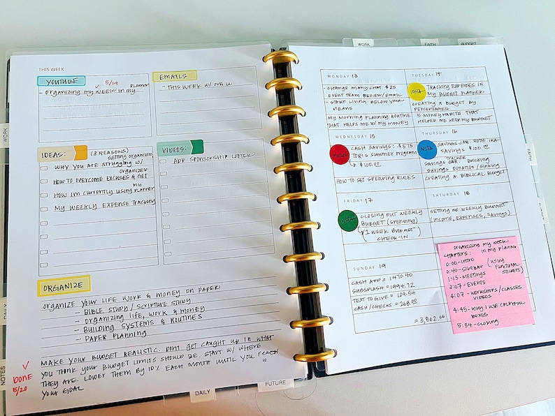 May include: A weekly planner with a black and white cover and gold rings. The planner is open to a page with a weekly schedule and a page with notes and ideas. The notes include "organize your life, work, and money on paper" and "make your budget realistic".