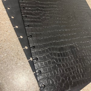 May include: Two black sheets of paper with a crocodile skin pattern. The sheets are joined together by a series of metal rings.