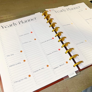 Op de afbeelding: Een jaarplanner met een gouden spiraalbinding en een transparante plastic hoes. De planner heeft een witte achtergrond met zwarte lijnen en handschrift. De tekst omvat "Yearly Planner", "Tax Day", "Easter Sunday", "Memorial Day", "Family Fun Day", "Women's Brunch" en "FUTURE".