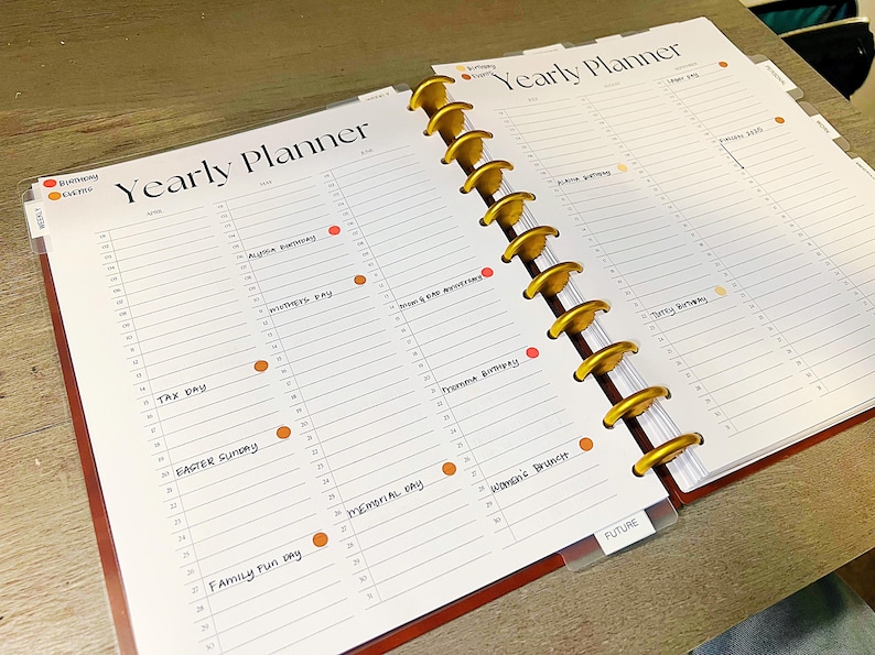 Op de afbeelding: Een jaarplanner met een gouden spiraalbinding en een transparante plastic hoes. De planner heeft een witte achtergrond met zwarte lijnen en handschrift. De tekst omvat "Yearly Planner", "Tax Day", "Easter Sunday", "Memorial Day", "Family Fun Day", "Women's Brunch" en "FUTURE".