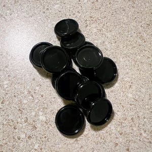 May include: A collection of glossy black, circular objects, possibly binder discs, are scattered across a speckled, light-colored surface. The discs are uniform in size and shape, with a slightly concave center.