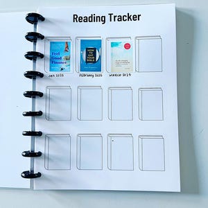 Könnte beinhalten: Eine weiße "Reading Tracker" Planerseite mit schwarzer Spiralbindung. Die Seite zeigt ein Raster von Buchumrissen, mit drei ausgefüllten Buchumschlägen, die mit Januar, Februar und März 2025 beschriftet sind. Das erste Buch ist "Feel Good Finance".