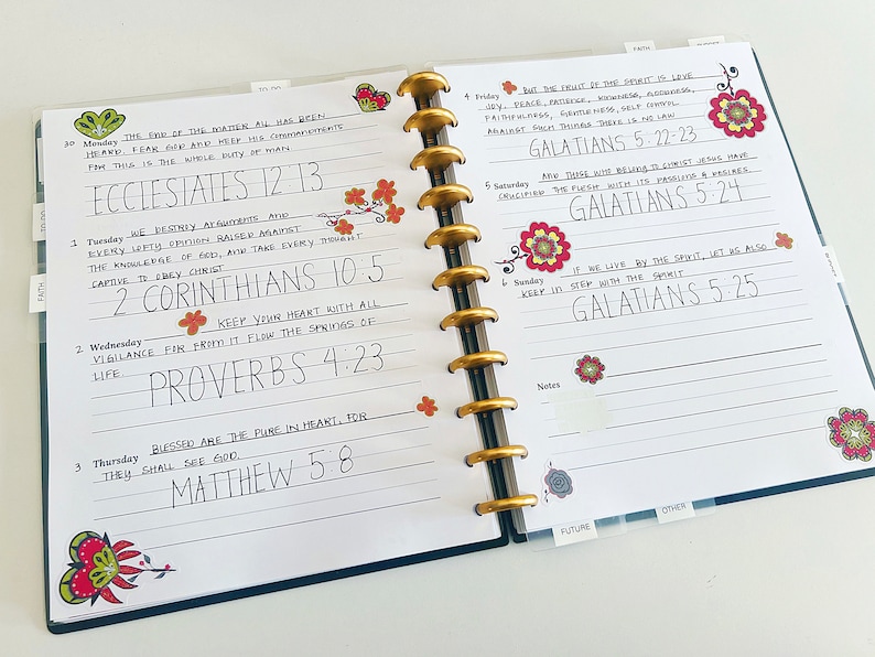May include: A weekly planner with a blue cover and white pages. The pages are filled with handwritten notes and bible verses. The planner has a gold coil binding and is decorated with colorful floral stickers.