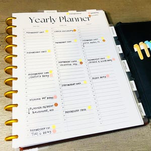 Op de afbeelding: Een witte jaarplanner met gouden ringen en een zwarte omslag. De planner heeft een sectie voor elke maand van het jaar en bevat een sectie voor persoonlijke afspraken, vergaderingen, geloof en budget. De planner is open op de februaripagina en heeft verschillende gekleurde stippen en stickers die belangrijke data markeren.