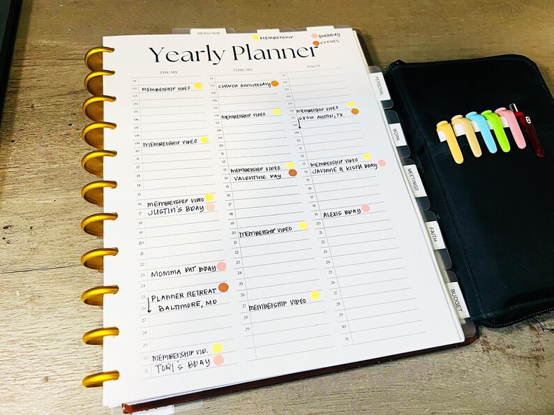 Op de afbeelding: Een witte jaarplanner met gouden ringen en een zwarte omslag. De planner heeft een sectie voor elke maand van het jaar en bevat een sectie voor persoonlijke afspraken, vergaderingen, geloof en budget. De planner is open op de februaripagina en heeft verschillende gekleurde stippen en stickers die belangrijke data markeren.