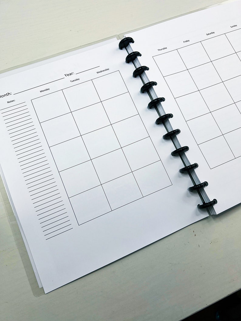 May include: A white planner with a black coil binding. The planner has a monthly calendar layout with spaces for notes and a section for the year and month.