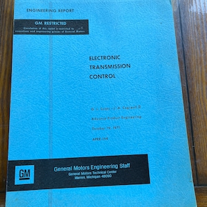 May include: A blue cover of an engineering report titled "Electronic Transmission Control" by General Motors Engineering Staff. The report was written by G. L. Casey and J. A. Cogswell II and published on October 19, 1971. The report number is APER-144.
