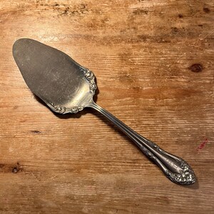 May include: A silver cake server with an ornate design on the handle. The server is lying on a wooden surface.