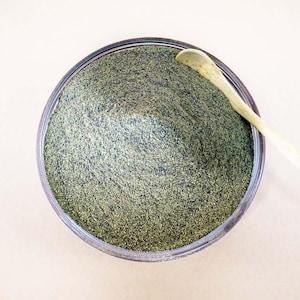 May include: A clear glass bowl filled with finely ground green powder, possibly a spice or herb. A small, light-colored wooden spoon rests on the edge of the bowl. The background is a neutral, off-white color.