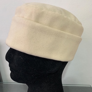 Può includere: Un cappello di feltro bianco con una corona arrotondata e un bordo ripiegato. Il cappello è su una testa di manichino nera.