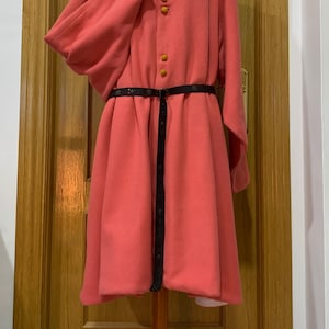 Può includere: Un lungo cappotto di lana rosa con collo alto e cintura nera. Il cappotto ha una fila di bottoni arancioni sul davanti.