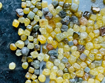 Puede incluir: Primer plano de una pequeña pala de metal llena de una gran cantidad de pequeños diamantes amarillos y grises rugosos. Los diamantes están dispersos sobre una superficie oscura.