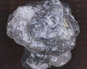 Puede incluir: Un diamante en bruto, sin cortar, con una estructura cristalina. La gema incolora tiene un aspecto nublado y una superficie texturizada, que refleja la luz. La forma irregular y las facetas del diamante son visibles en esta toma de cerca.