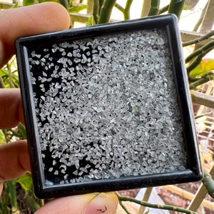 Puede incluir: Un marco cuadrado negro lleno de pequeños cristales de cuarzo transparentes. Los cristales están densamente empaquetados, creando un efecto brillante.