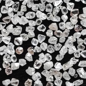 May include: A close-up view of a collection of rough, uncut diamonds. The diamonds are various sizes and shapes, with some having a slightly brownish hue.