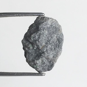 May include: A rough, dark gray, and green gemstone with a slightly oval shape. The gemstone is being held by a pair of tweezers.