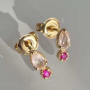 Op de afbeelding: Een paar gouden oorstekers. Elke oorbel heeft een peervormige, lichtroze edelsteen boven een kleine, ronde, felroze edelsteen. De oorbellen hebben een delicate, getextureerde gouden rand.